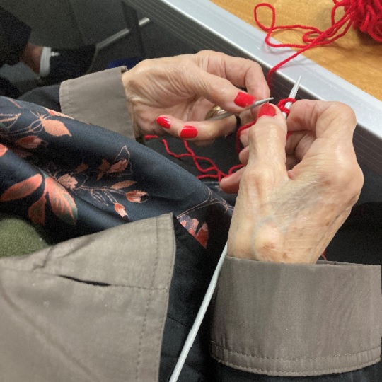 Photo of Maria's 91 year old hands with bright red nail polish knitting with red wool, her coat and scarf are visible in the photo